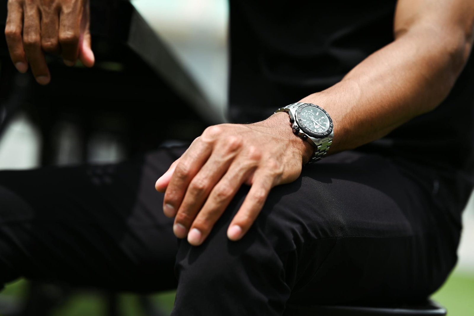 Close-up image featuring a man's hands with a silver wristwatch, wearing black pants.